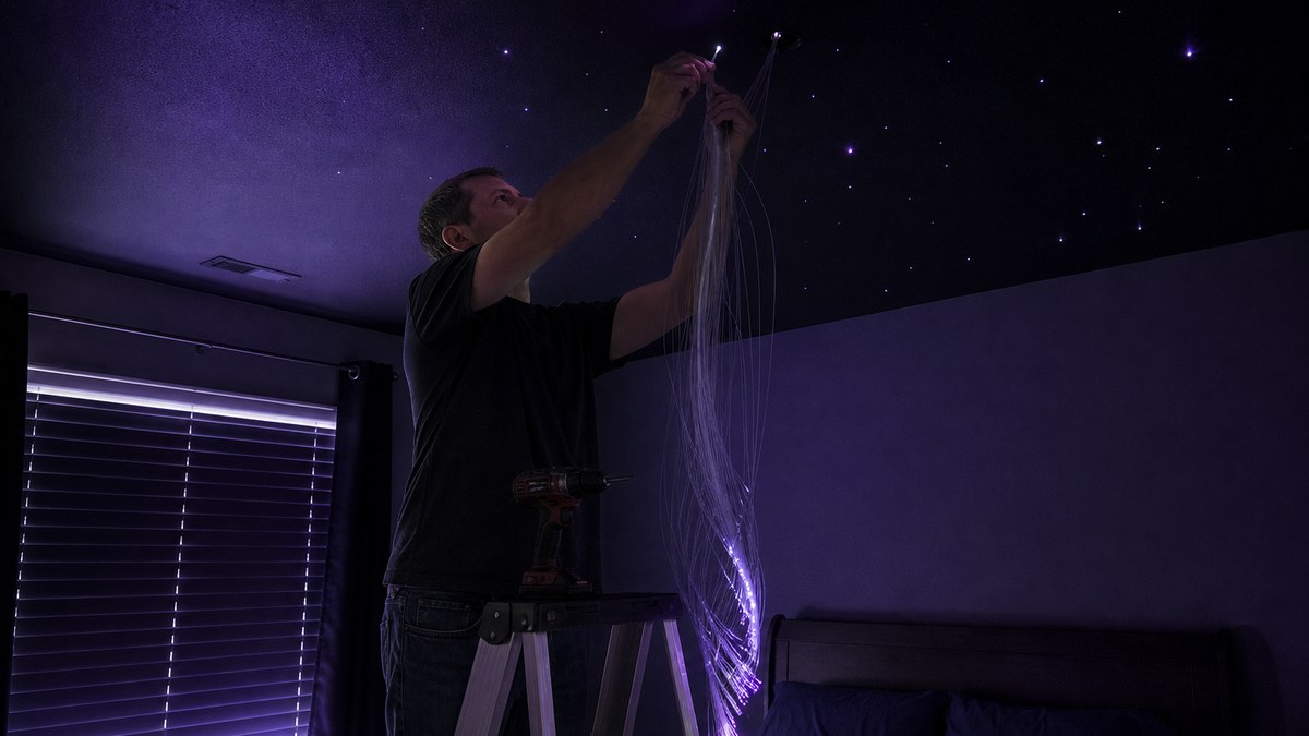 DIY star ceiling installation in progress with a homeowner on a ladder threading fiber optic strands through drilled holes in drywall