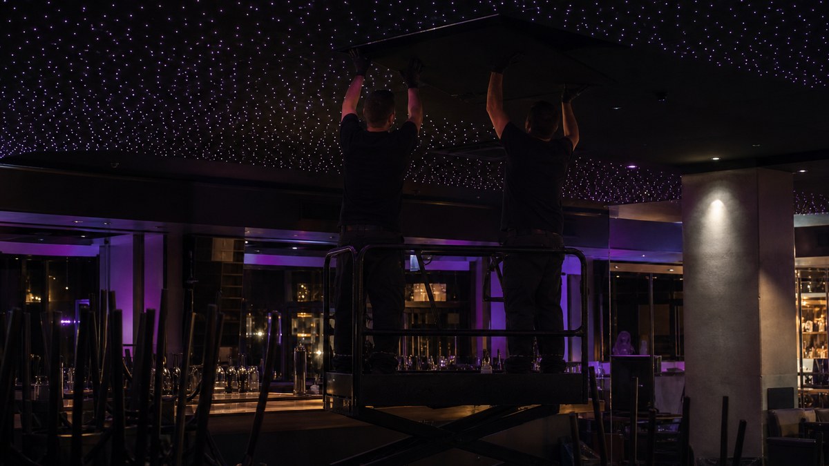 Two installers on a scissor lift mounting fiber optic star ceiling panels in a closed restaurant bar after hours